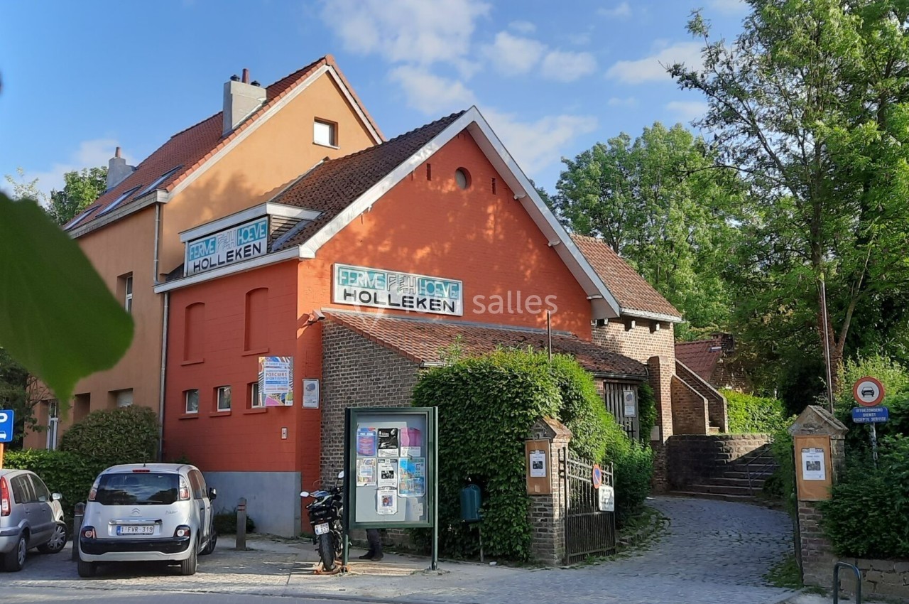 Location salle Linkebeek (Brabant flamand) - Ferme Holleken Hoeve #7 Bâtiment en briques rouges avec des panneaux ’Holleken’, entouré de végétation, voitures garées et panneau d'affichage.