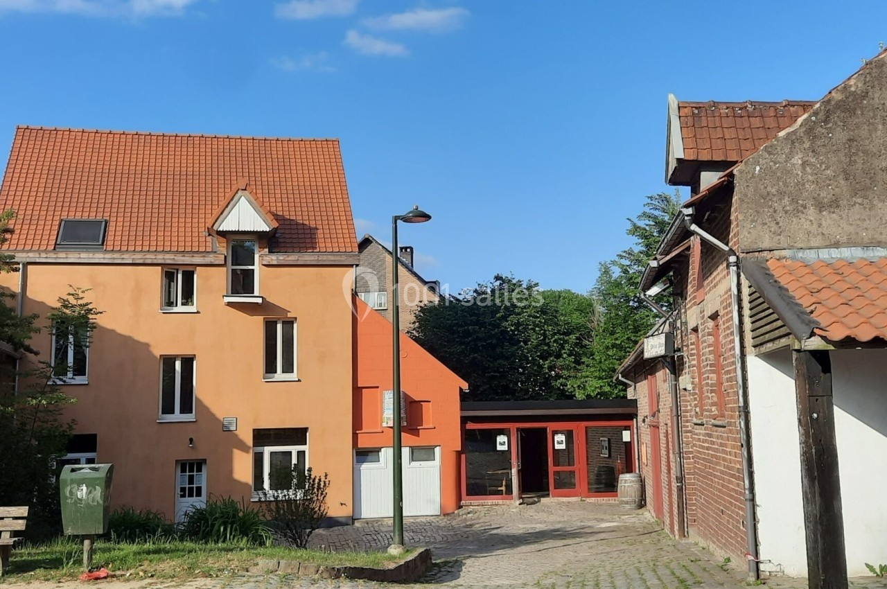 Location salle Linkebeek (Brabant flamand) - Ferme Holleken Hoeve #8 Maisons en briques et crépi orange avec toits en tuiles, bordant une ruelle pavée sous un ciel bleu.