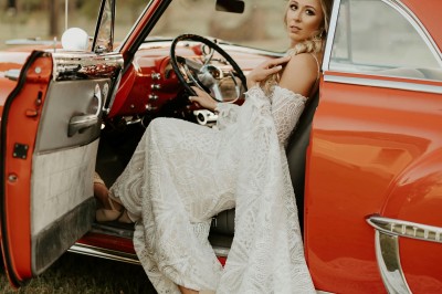 Une femme en robe de mariée tient un bouquet de tournesols près d'une voiture ancienne rouge, de nuit.