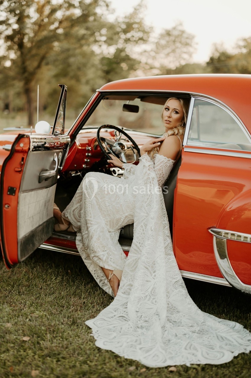 Une femme en robe de mariée blanche est assise dans une voiture ancienne rouge, porte ouverte, dans un cadre extérieur.