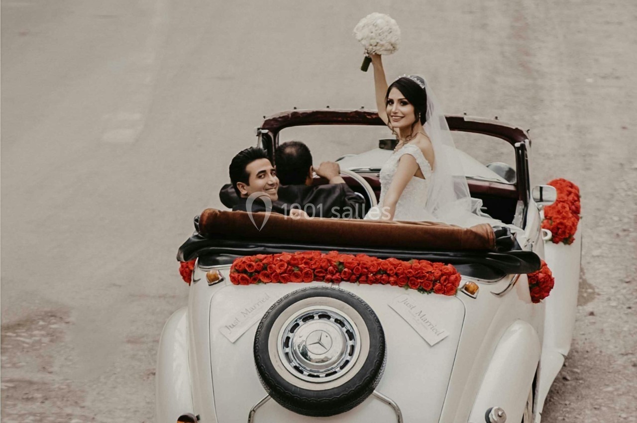 Un couple de mariés souriants dans une voiture ancienne décorée de roses rouges, la mariée levant un bouquet.