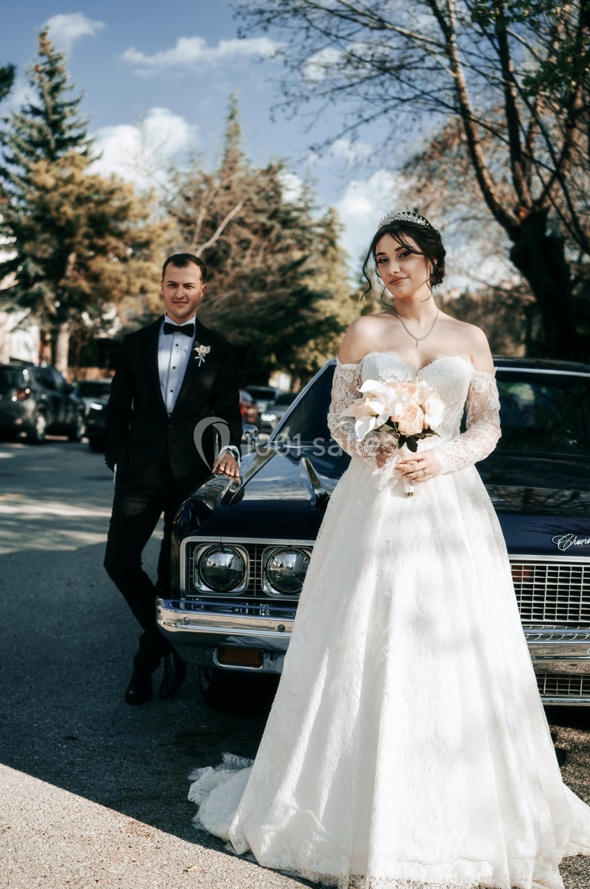 Une mariée en robe blanche tient un bouquet devant une voiture ancienne, un homme en costume se tient à côté.