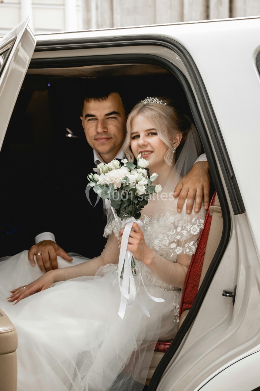 Un couple de mariés souriant assis dans une voiture, la mariée tenant un bouquet de fleurs blanches.