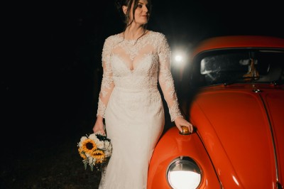 Une femme en robe de mariée tient un bouquet de tournesols près d'une voiture ancienne rouge, de nuit.