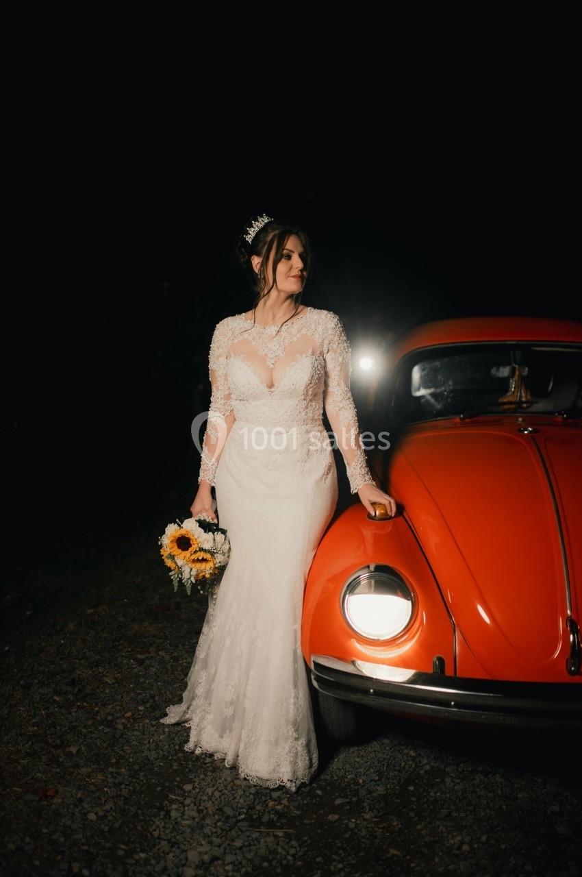 Une femme en robe de mariée tient un bouquet de tournesols près d'une voiture ancienne rouge, de nuit.