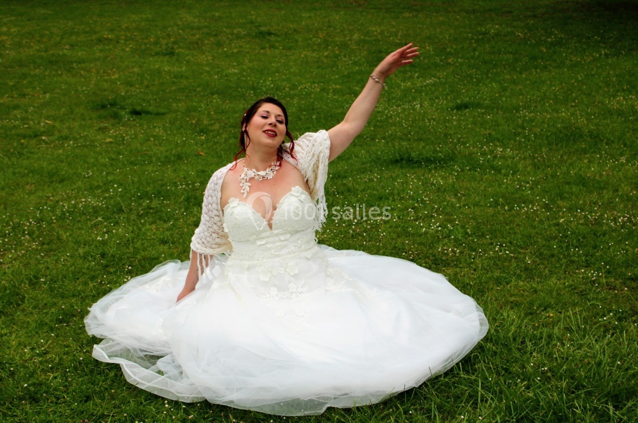 Femme en robe de mariée blanche assise sur une pelouse verte, levant un bras avec une expression détendue.