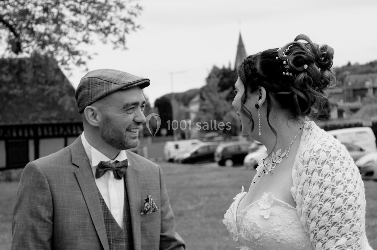Un couple en tenue de mariage échange un sourire dans un cadre extérieur avec des bâtiments en arrière-plan.