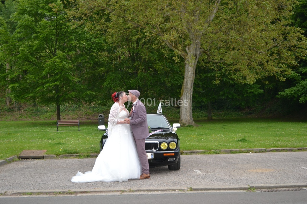 Un couple en tenue de mariage s'embrasse devant une voiture noire, dans un parc verdoyant.