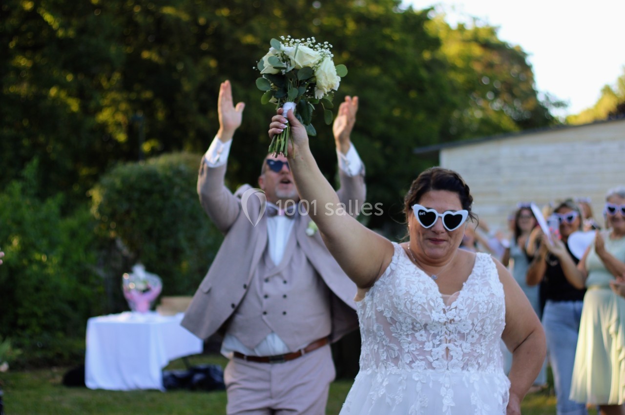 Une mariée souriante portant des lunettes en forme de cœur lève son bouquet, entourée d'invités dans un jardin.