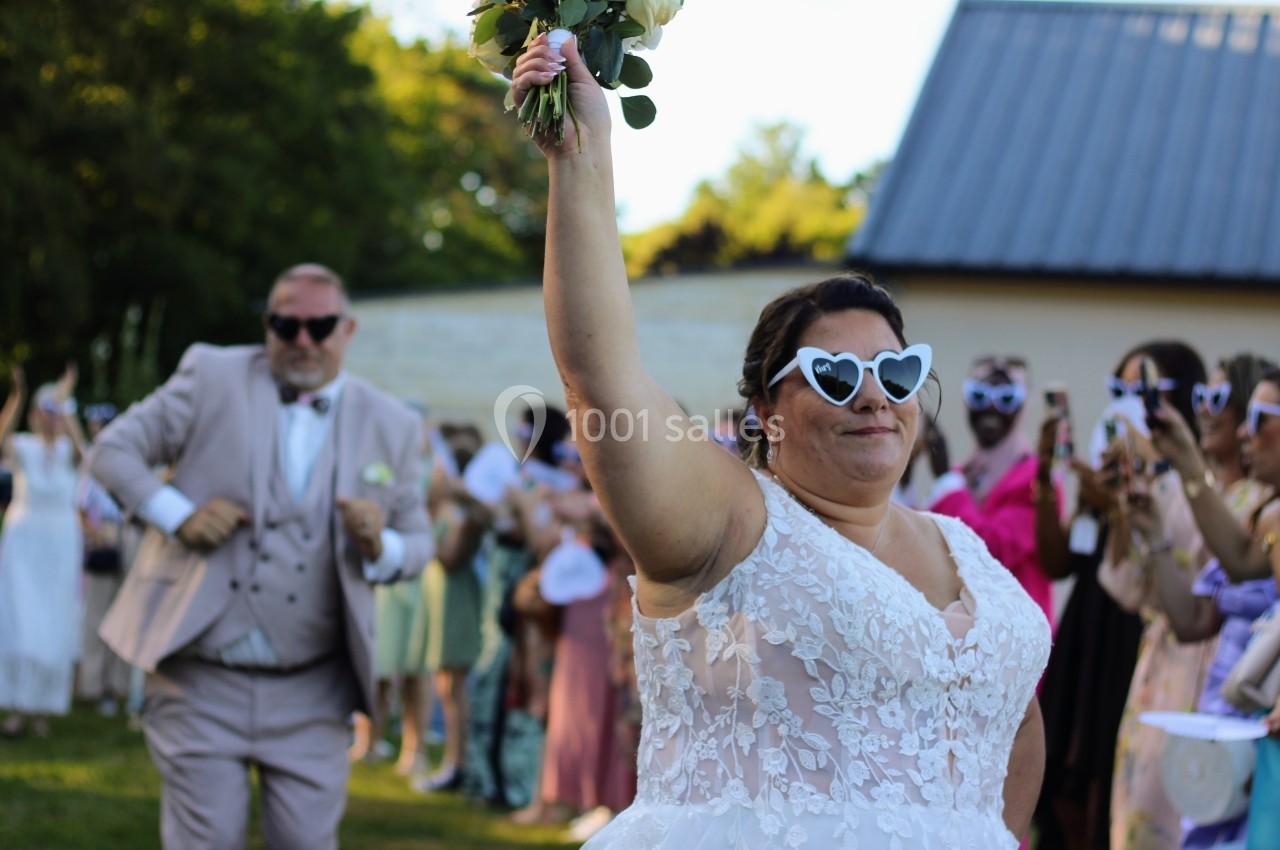 Une mariée en robe blanche et lunettes en forme de cœur lève un bouquet, entourée d'invités lors d'une fête en extérieur.