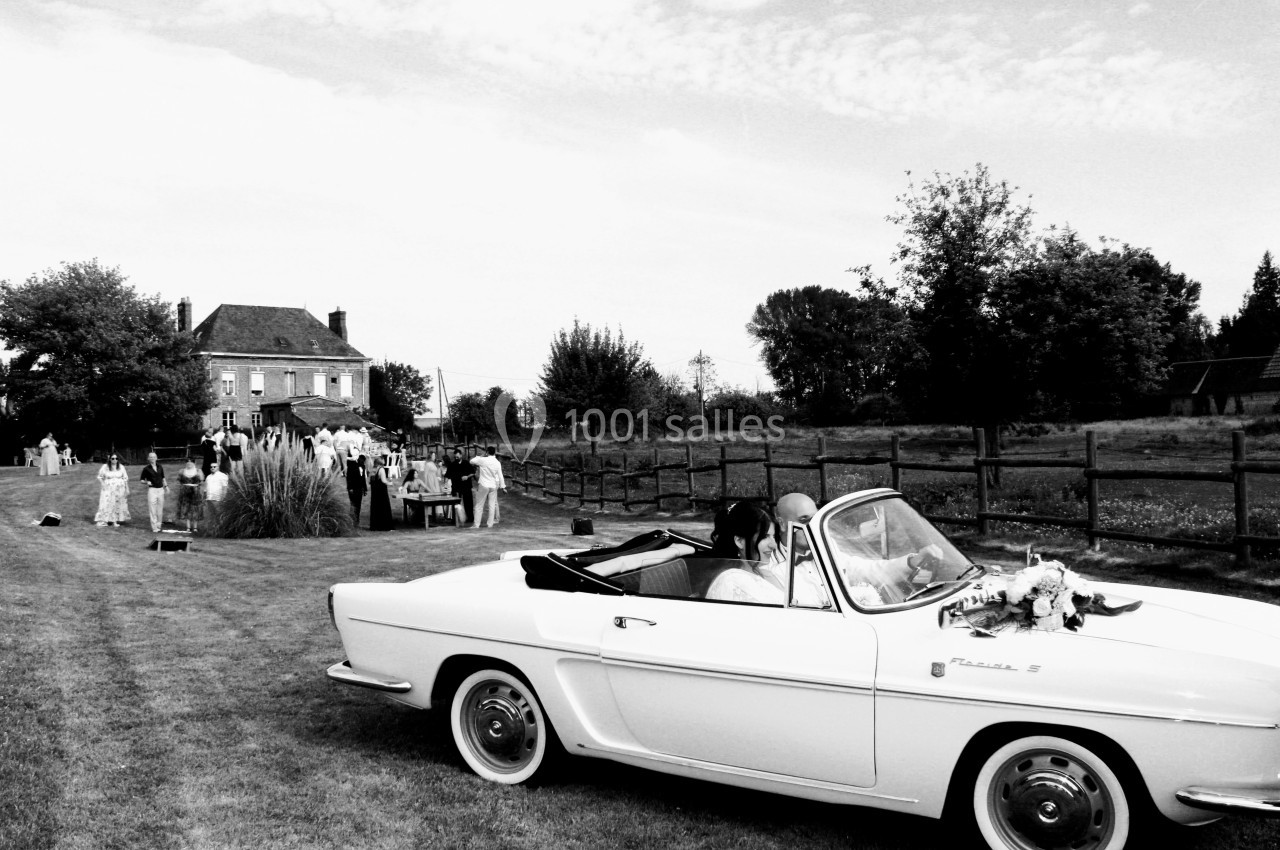 Voiture décapotable blanche garée sur une pelouse, avec des invités rassemblés près d'une maison en arrière-plan.