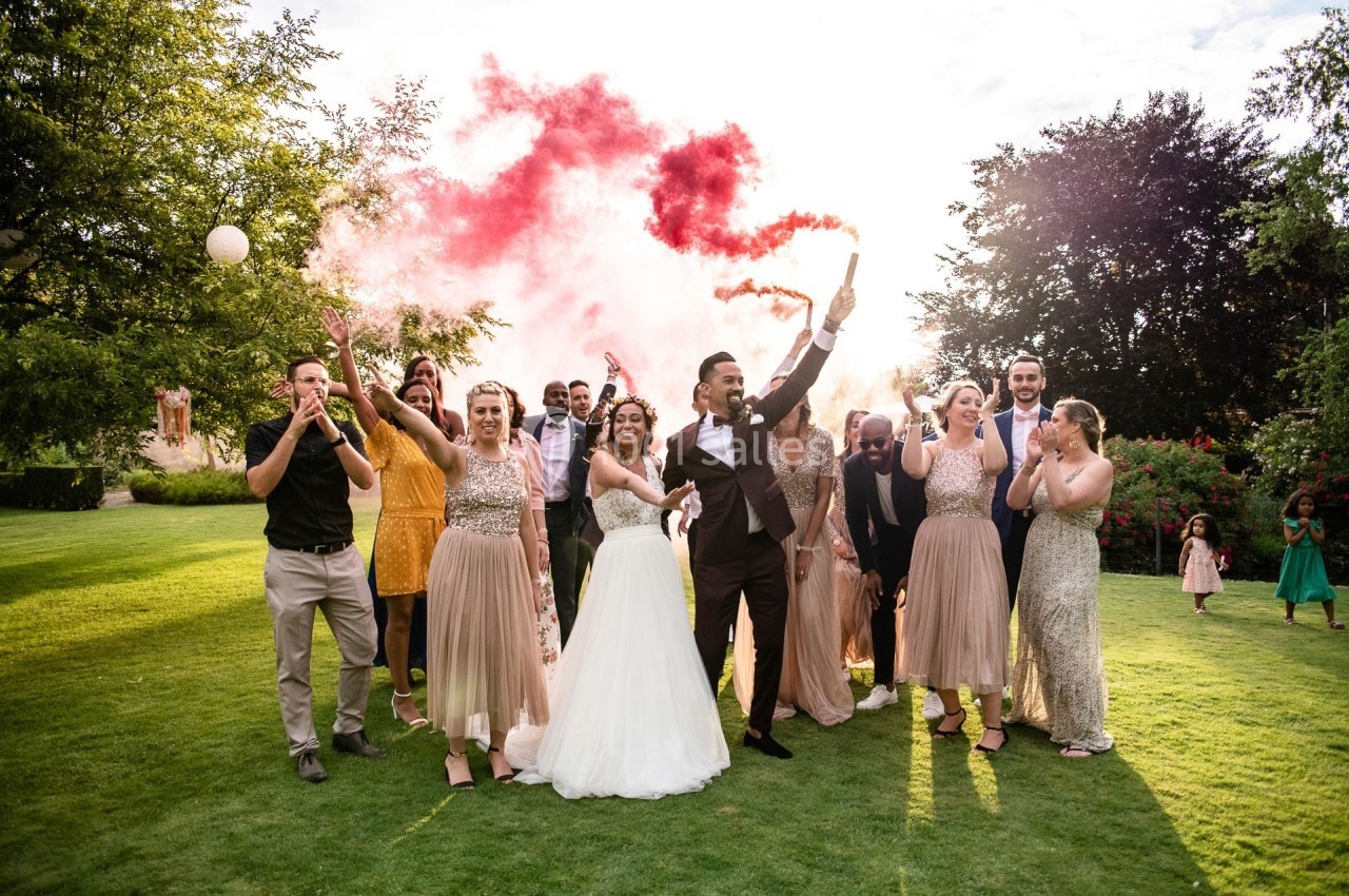 Un couple de mariés entouré d'invités célébrant avec des fumigènes colorés dans un jardin ensoleillé.