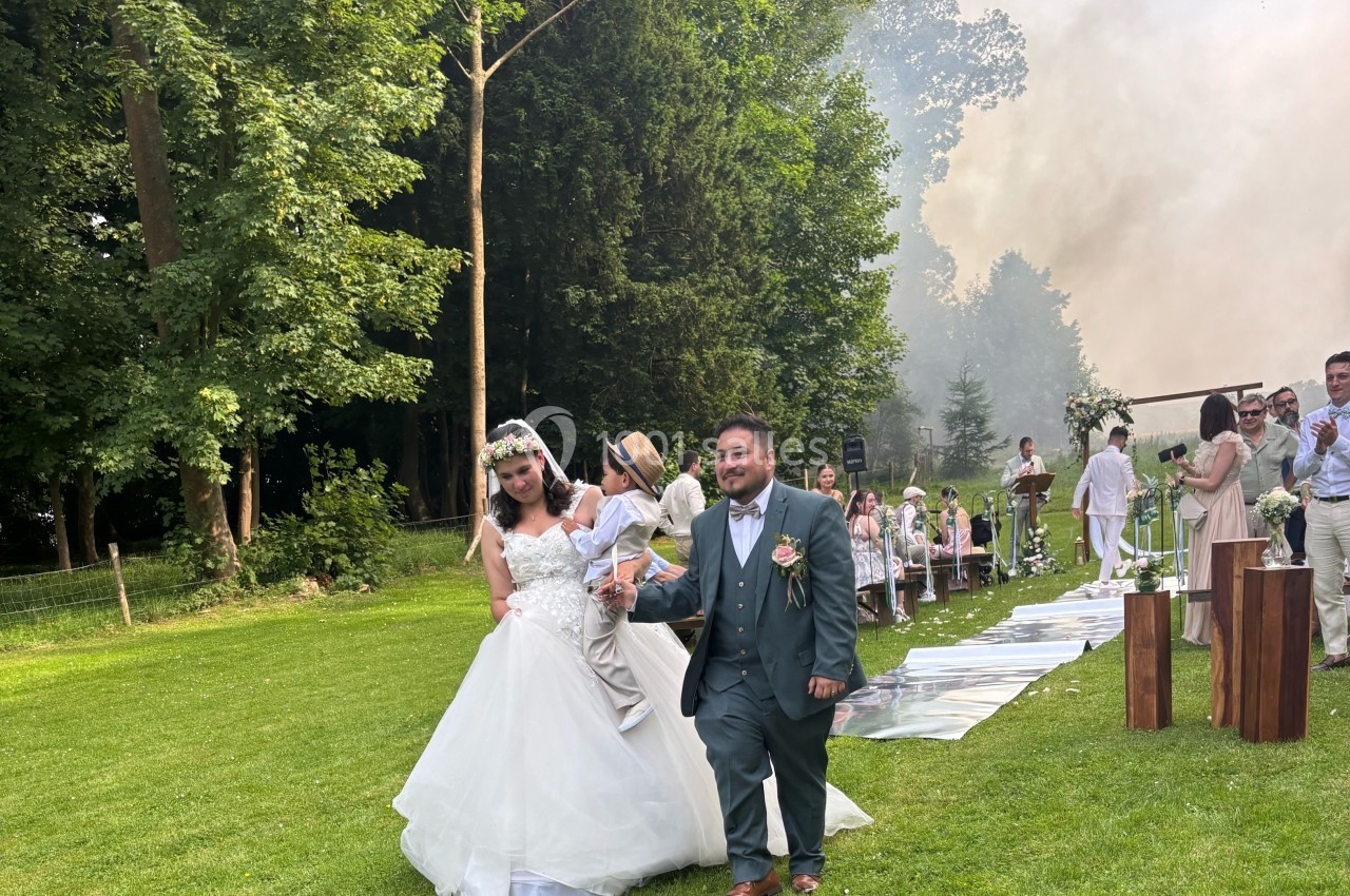 Un couple de mariés marche sur une pelouse, entouré d'invités et d'arbres, lors d'une cérémonie en plein air.