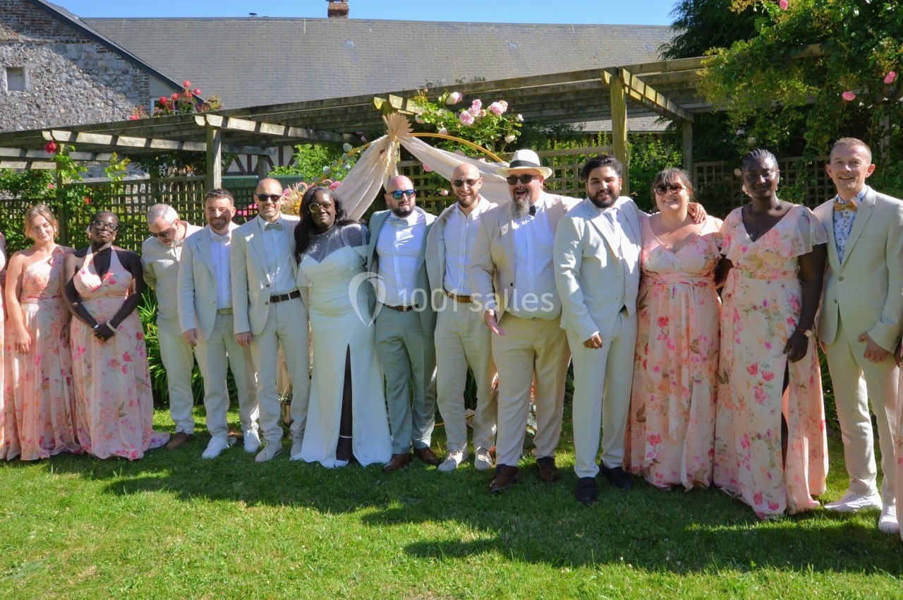 Un groupe de personnes en tenue de mariage pose dans un jardin ensoleillé devant une pergola fleurie.