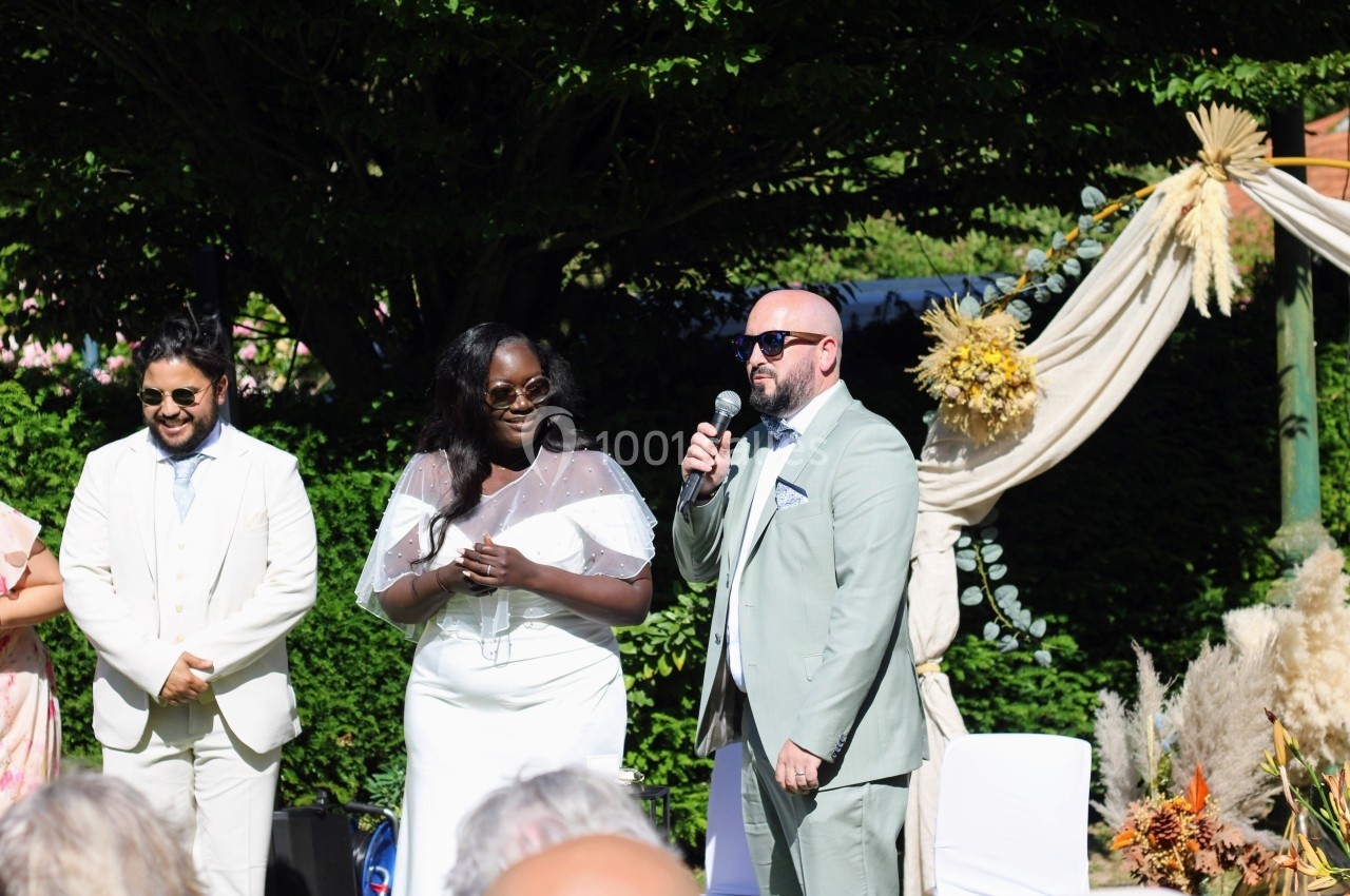 Un couple en tenue de mariage se tient debout à l'extérieur, accompagné d'un homme en costume, sous une arche décorée.