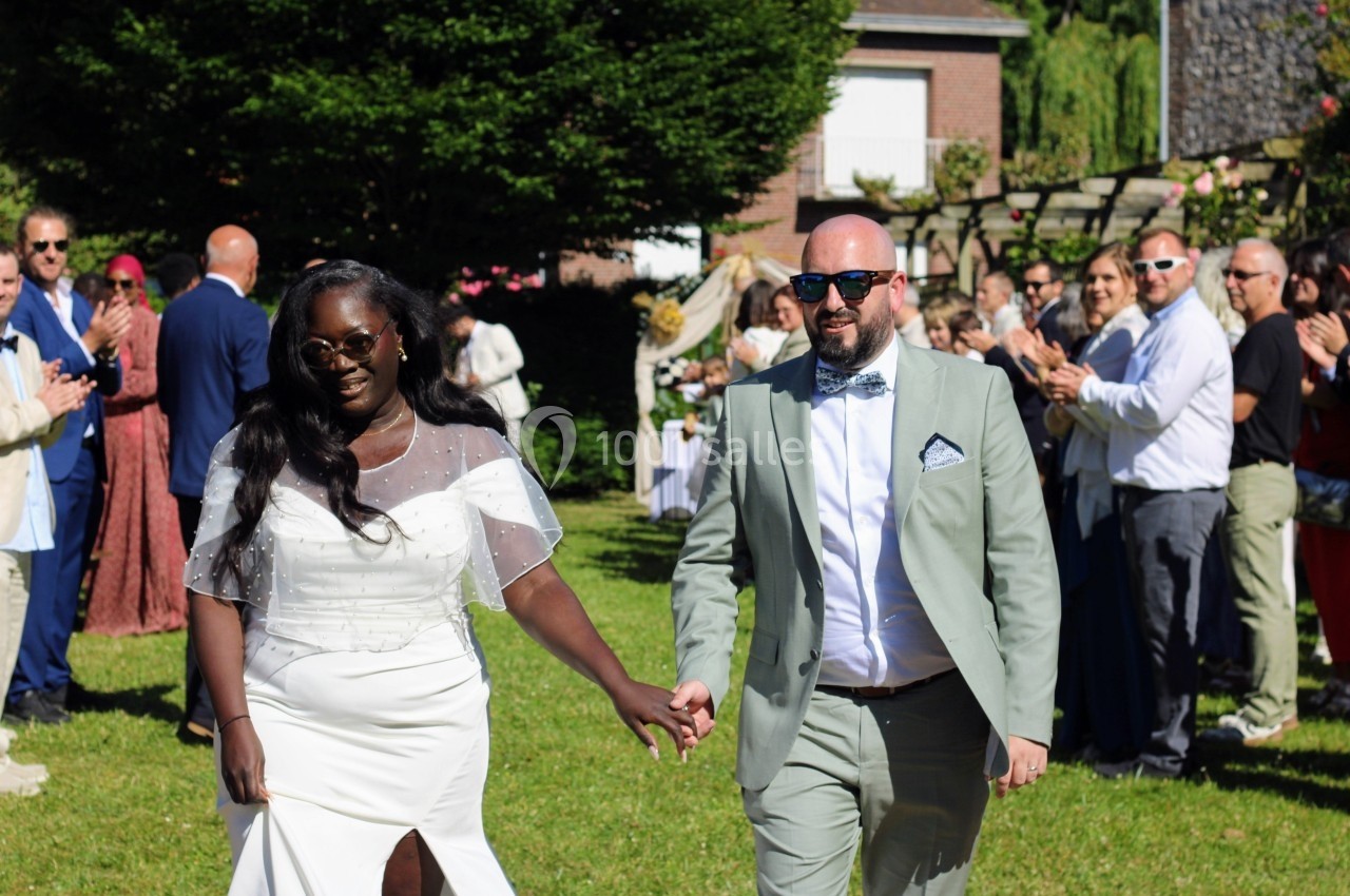 Un couple souriant marche main dans la main dans un jardin, entouré d'invités applaudissant.