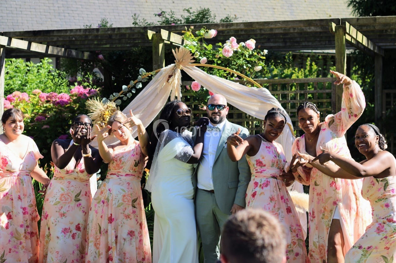 Un couple de mariés pose joyeusement avec des demoiselles d'honneur en robes fleuries dans un jardin ensoleillé.