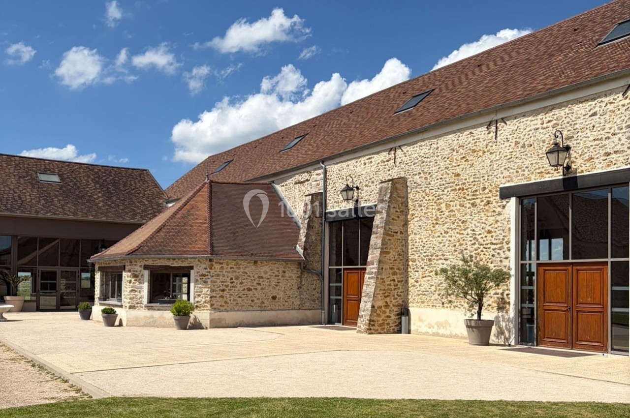 Bâtiment en pierre avec toit en tuiles, grandes portes en bois et espace extérieur pavé sous un ciel bleu.