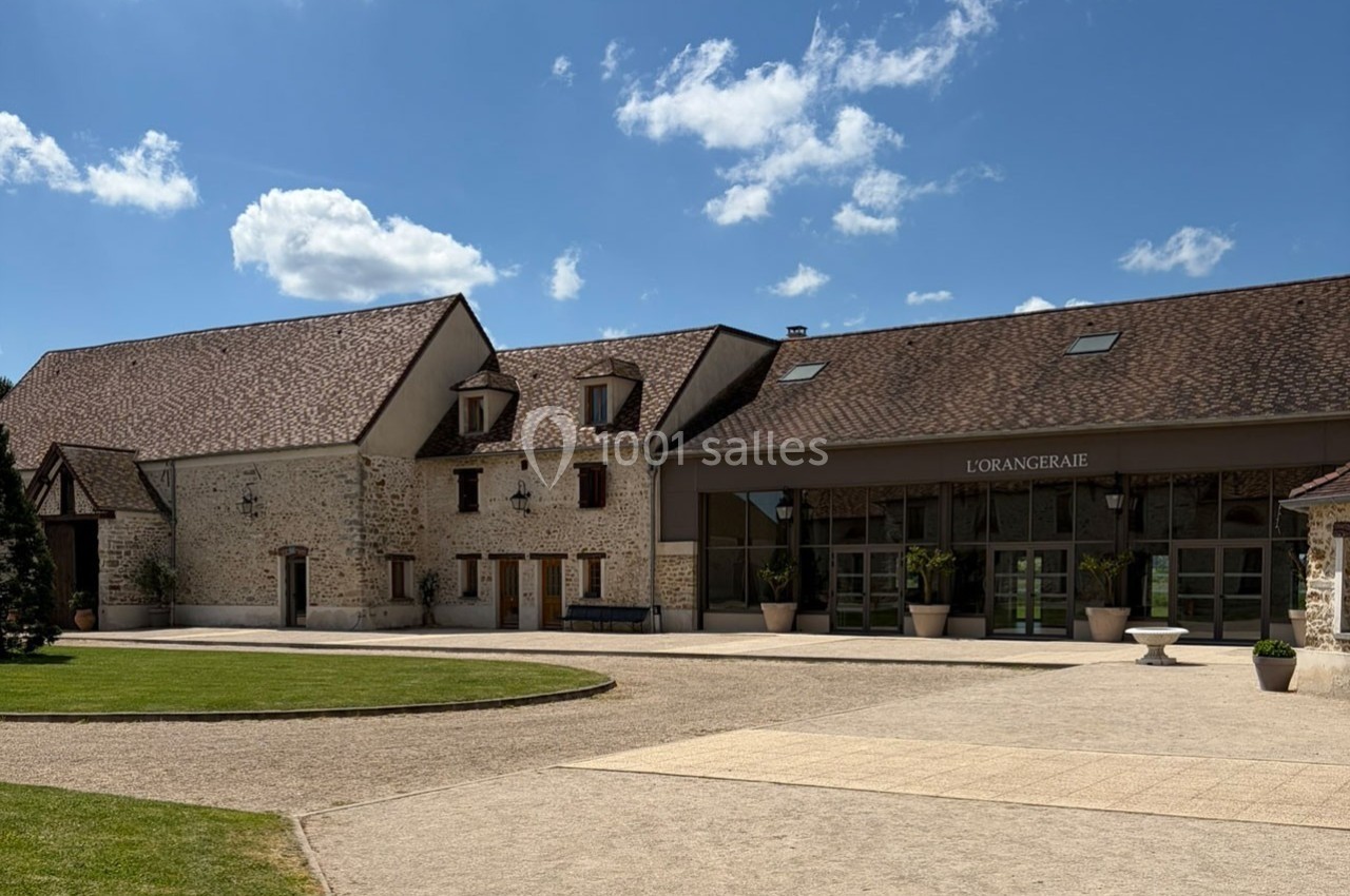 Bâtiment en pierre avec toit en tuiles, grande entrée vitrée et espace extérieur dégagé sous un ciel bleu.