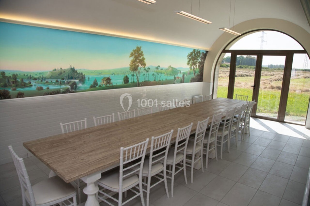 Grande table en bois entourée de chaises blanches dans une salle lumineuse avec fresque murale et vue sur un paysage…