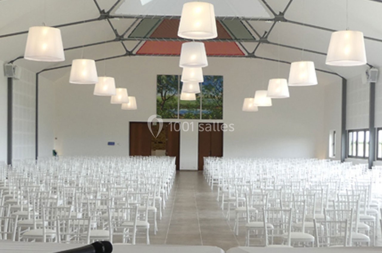 Salle de réception lumineuse avec des rangées de chaises blanches alignées sous des suspensions modernes.