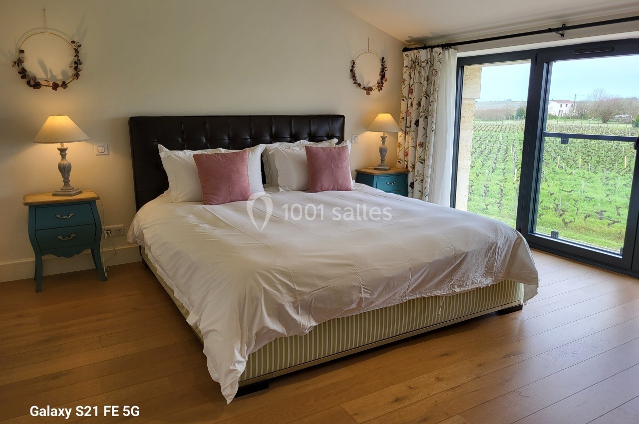 Chambre lumineuse avec lit double, tables de chevet, lampes et vue sur un vignoble par une grande baie vitrée.