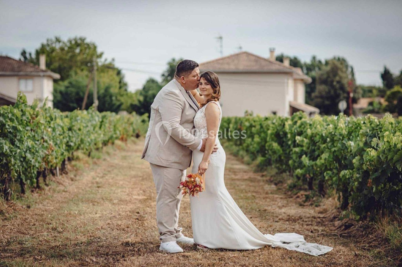 Un couple en tenue de mariage pose dans un vignoble, entouré de rangées de vignes sous un ciel clair.