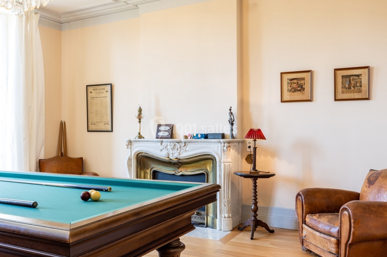 Salle avec une table de billard, cheminée décorative, fauteuils en cuir et cadres accrochés au mur beige.