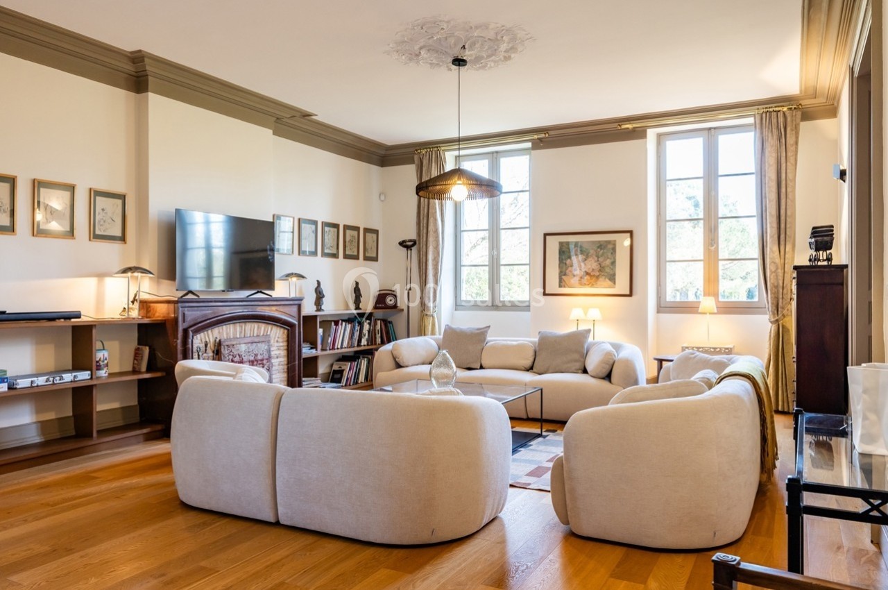 Salon lumineux avec parquet, canapés beiges, cheminée décorative, étagères et grandes fenêtres laissant entrer la lumière.