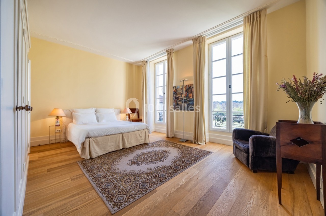 Chambre lumineuse avec lit double, tapis, fauteuil, bureau en bois et grandes fenêtres donnant sur un paysage.