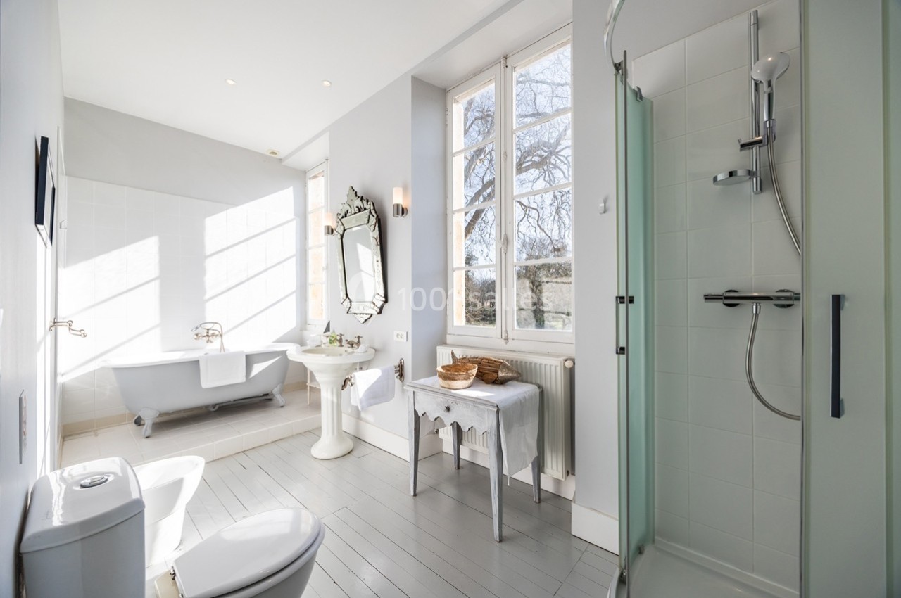 Salle de bain lumineuse avec baignoire sur pieds, douche vitrée, lavabo sur pied et grande fenêtre donnant sur l'extérieur.