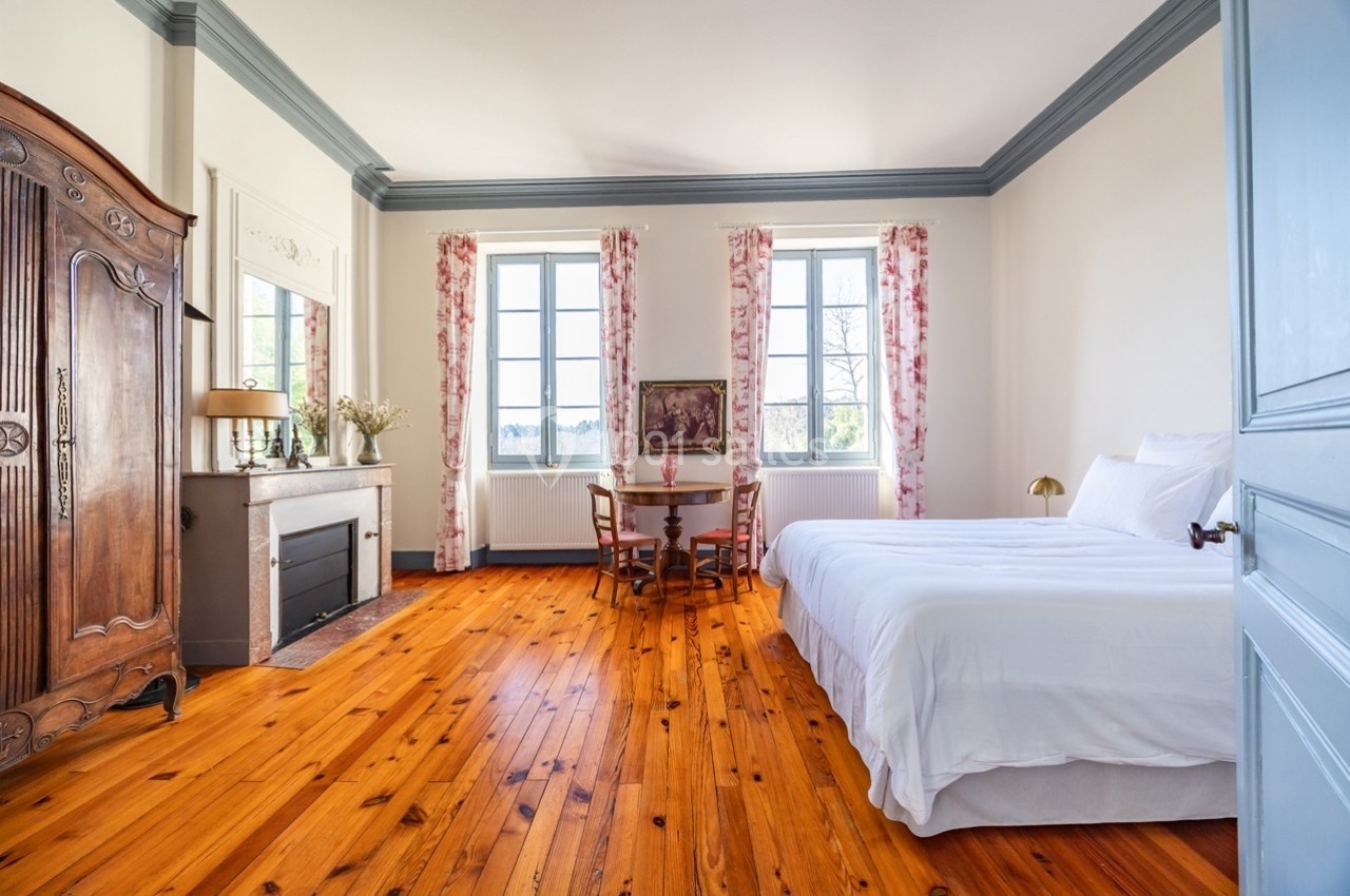 Chambre lumineuse avec parquet en bois, lit double, cheminée, armoire ancienne et table près de fenêtres avec rideaux…