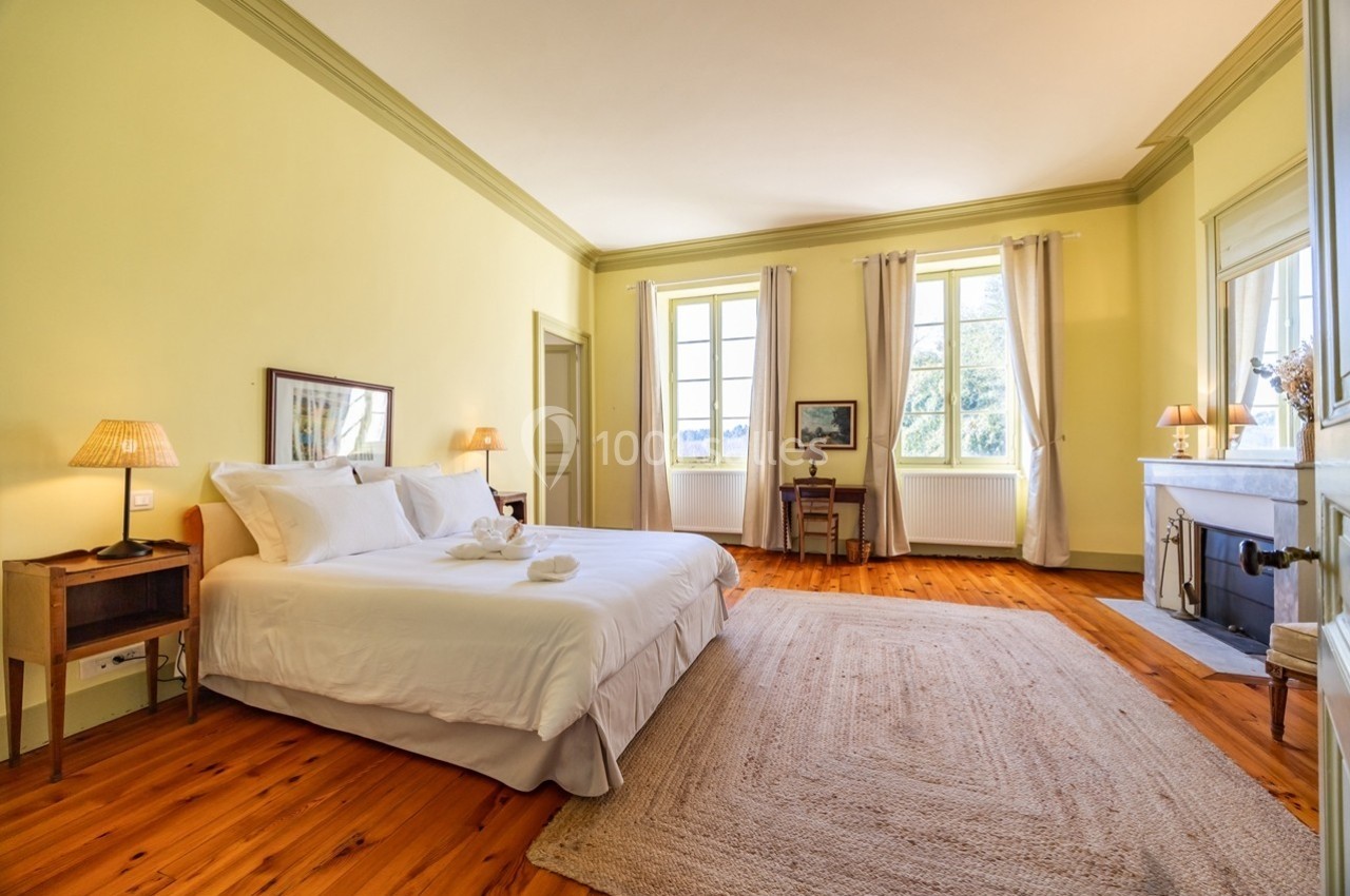 Chambre lumineuse avec lit double, parquet en bois, tapis beige, cheminée et grandes fenêtres donnant sur l'extérieur.