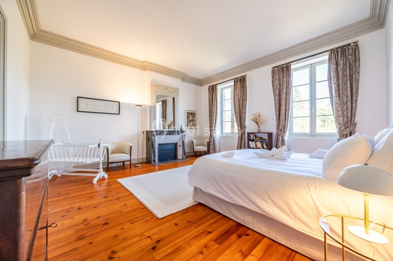 Chambre lumineuse avec lit double, parquet en bois, cheminée, berceau et grandes fenêtres avec rideaux.