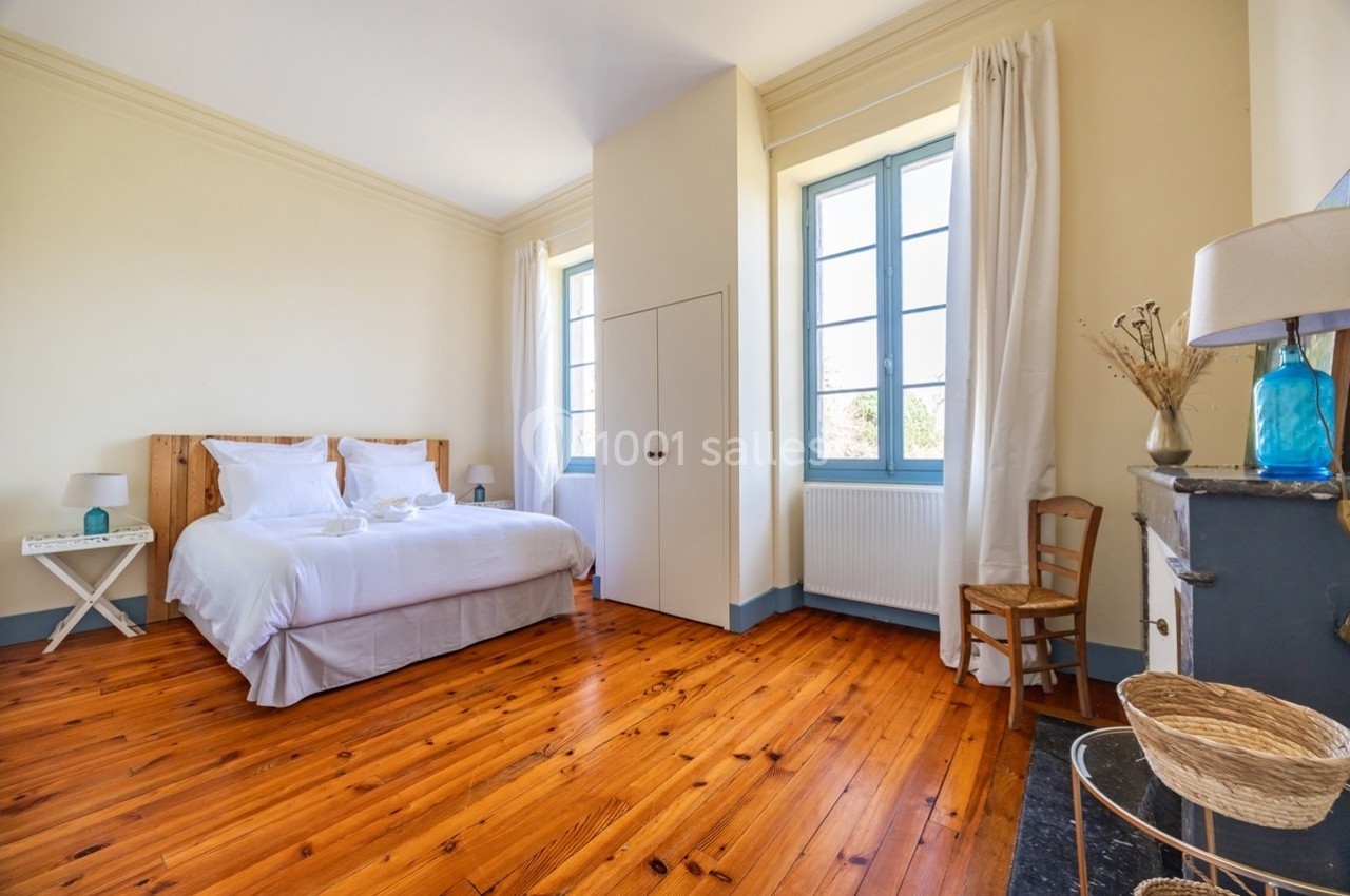 Chambre lumineuse avec lit double, parquet en bois, murs clairs, grandes fenêtres et mobilier minimaliste.