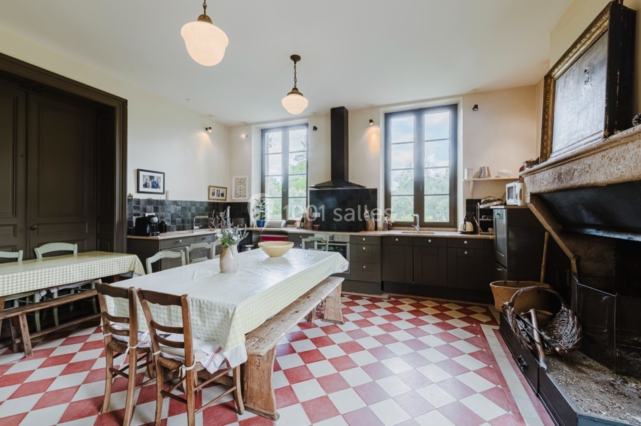 Cuisine rustique avec carrelage rouge et blanc, grande table en bois, cheminée ancienne et fenêtres lumineuses.