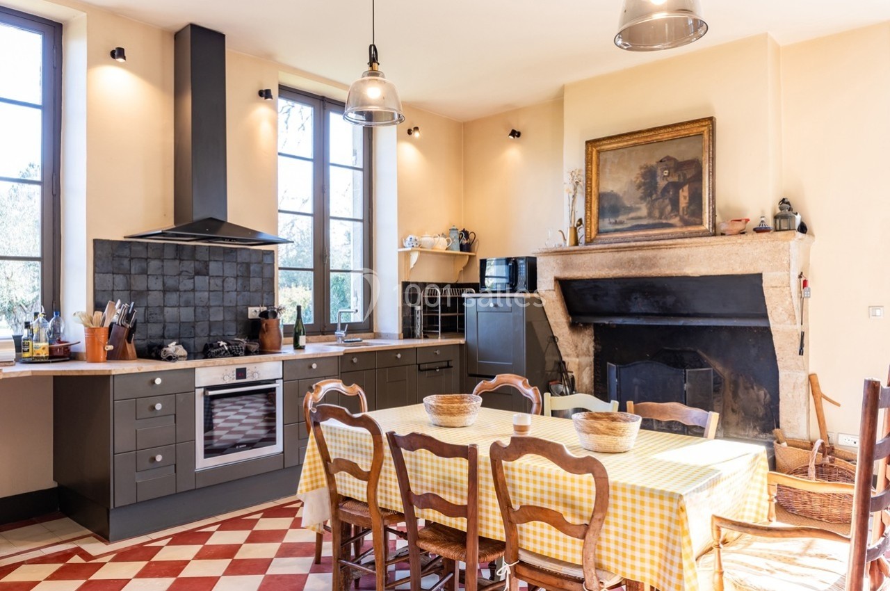 Cuisine lumineuse avec table en bois et nappe à carreaux, cheminée en pierre, meubles gris et grandes fenêtres.
