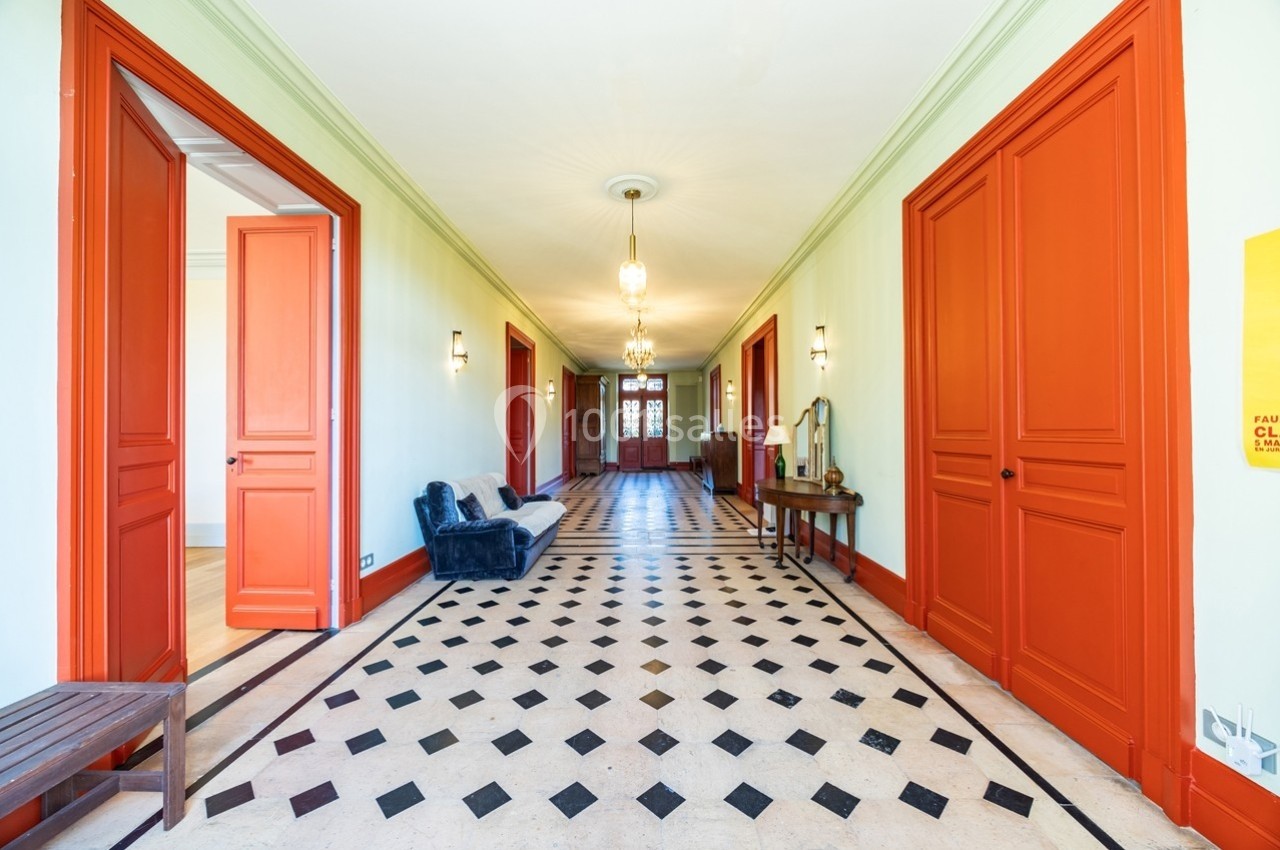 Couloir lumineux avec sol en damier, murs clairs, portes rouges et mobilier minimaliste.