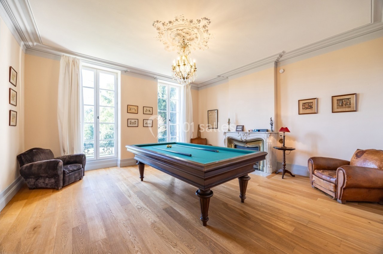 Salle lumineuse avec un billard au centre, parquet en bois, fauteuils et cheminée décorative.