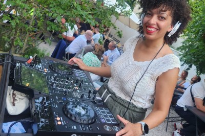 Miniature DJ Lyon (Rhône) - MetissKyla #4 Une DJ souriante mixe de la musique en plein air lors d'un événement, entourée de verdure et de participants.