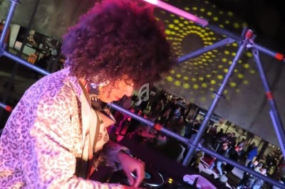 Une DJ mixe sur une table de mixage lors d'un événement en plein air, éclairée par des lumières colorées.