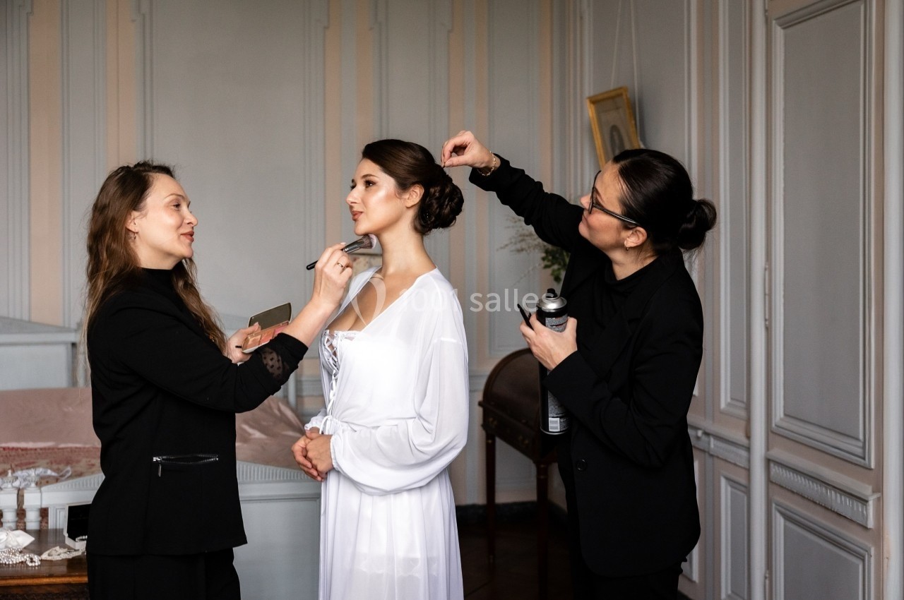 Deux maquilleuses préparent une femme en robe blanche dans une pièce lumineuse au décor classique.