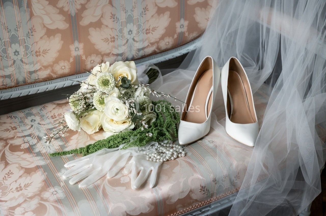 Bouquet de mariée, gants blancs, collier de perles et escarpins blancs posés sur un canapé fleuri avec un voile.