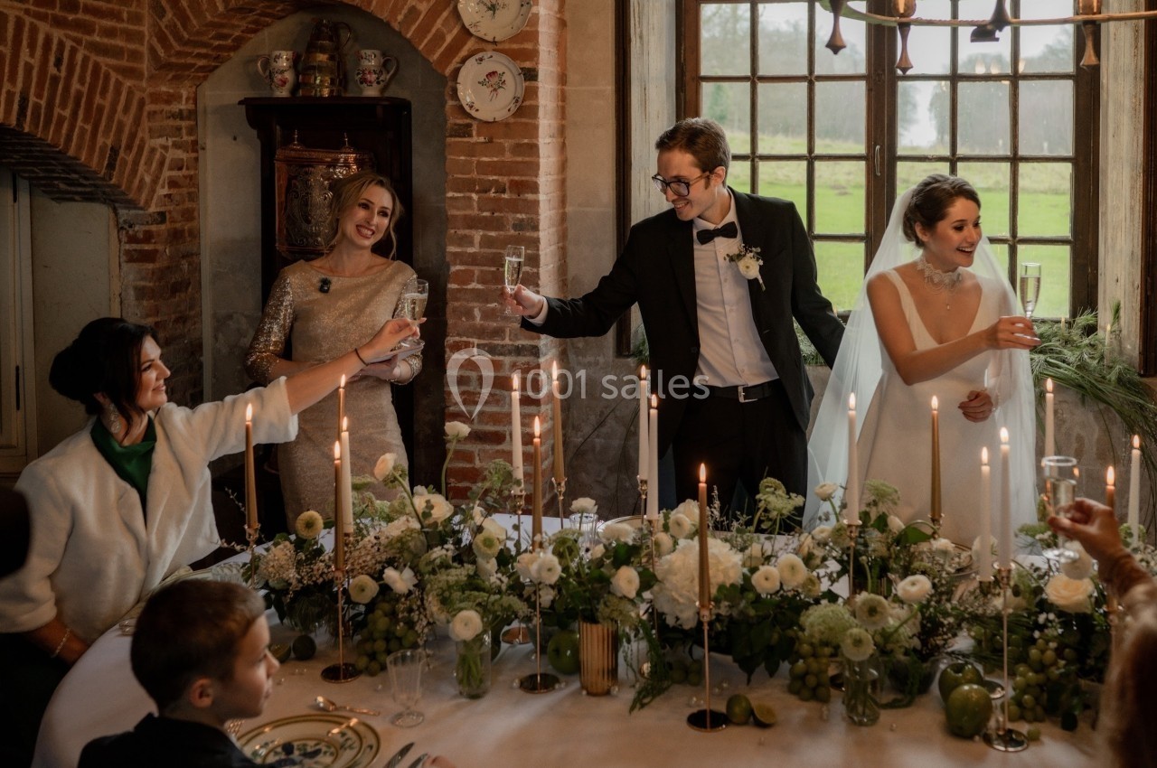 Des mariés et leurs invités trinquent autour d'une table décorée de fleurs et de bougies dans une salle rustique.