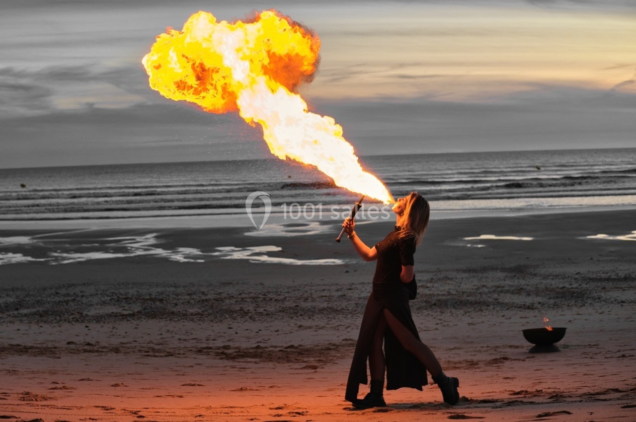 Une personne crache du feu sur une plage au coucher du soleil, avec une flamme vive illuminant la scène.