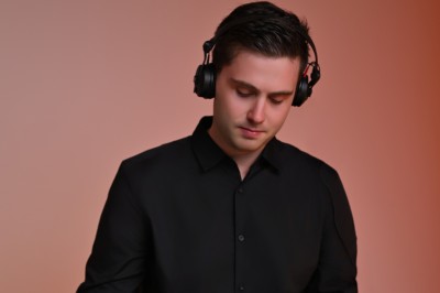 Un homme portant un casque audio noir, vêtu d'une chemise noire, se tient devant un fond dégradé orange.