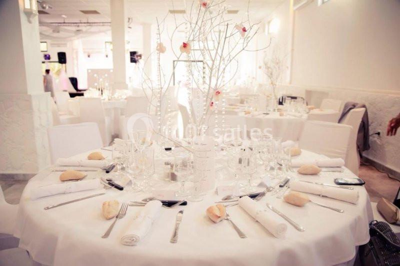 Table de réception élégamment dressée avec nappes blanches, couverts, verres, petits pains et centre de table décoratif.