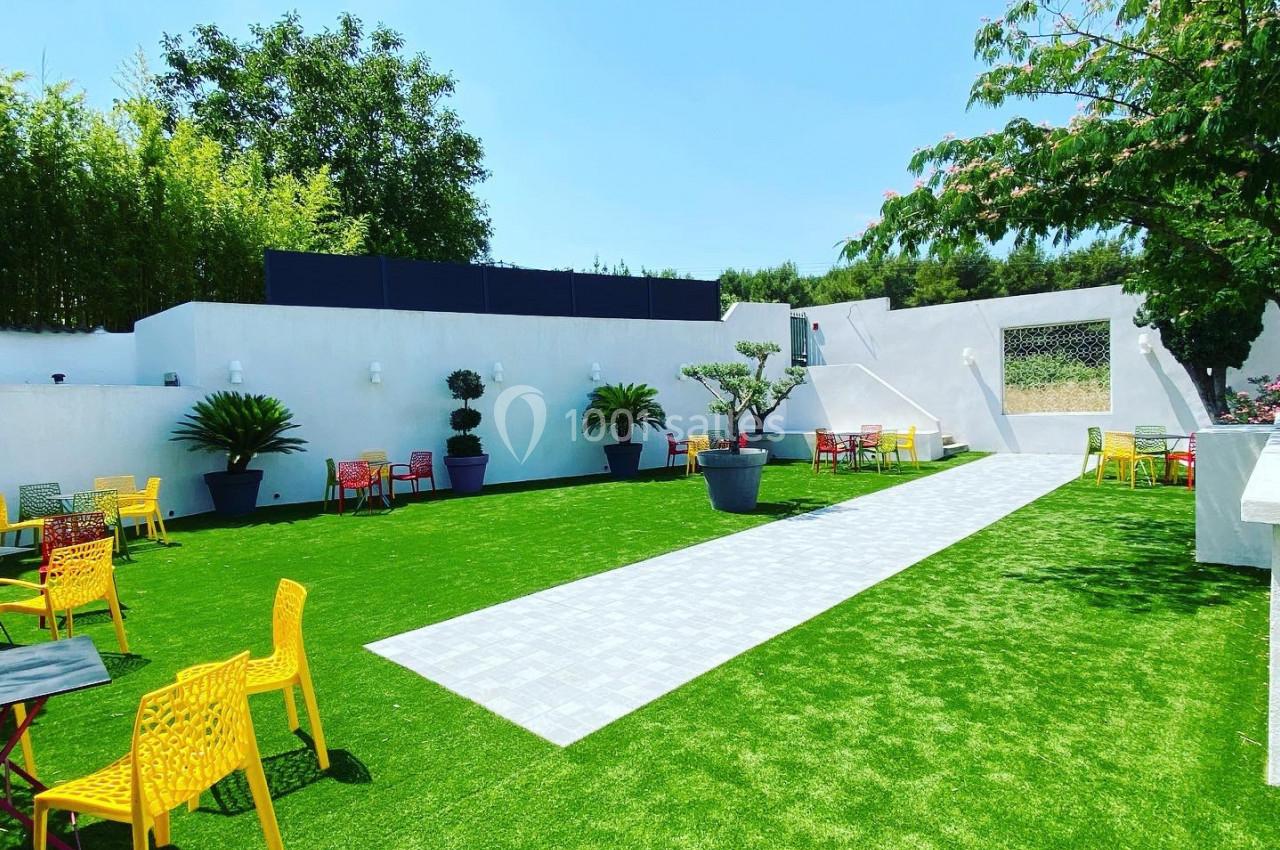 Cour extérieure avec pelouse artificielle, tables et chaises colorées, entourée de murs blancs et végétation.