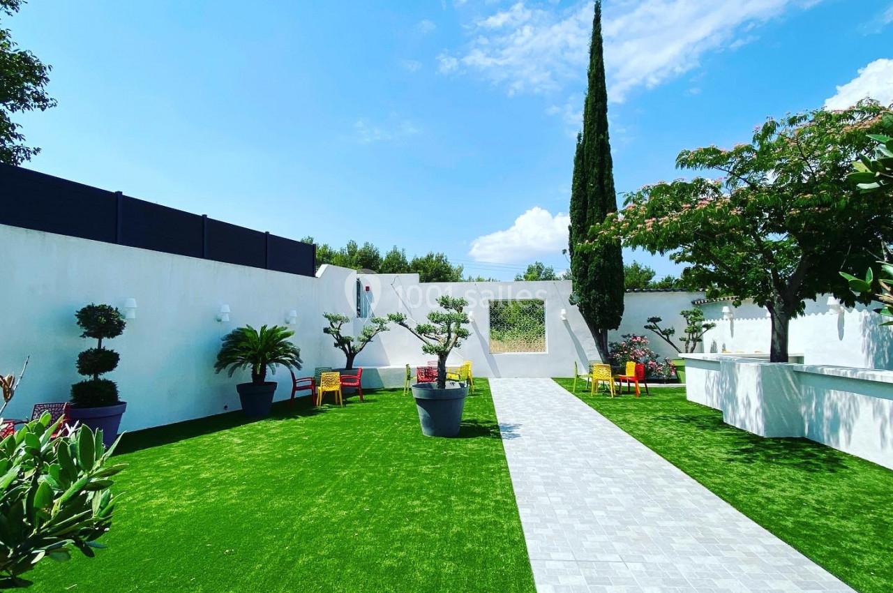 Jardin avec pelouse artificielle, allée pavée, plantes en pots et mobilier coloré sous un ciel bleu.