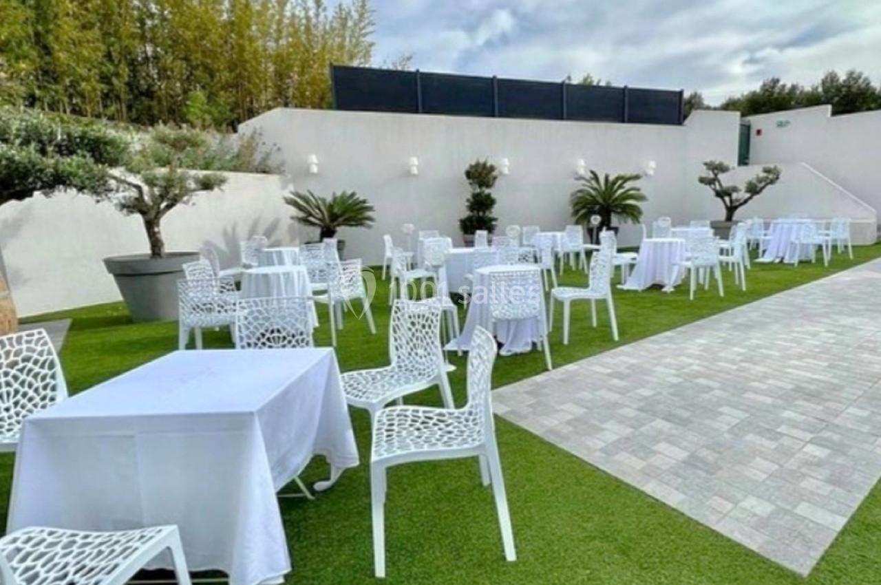 Tables et chaises blanches disposées sur une pelouse artificielle, entourées de plantes et d'un mur blanc.